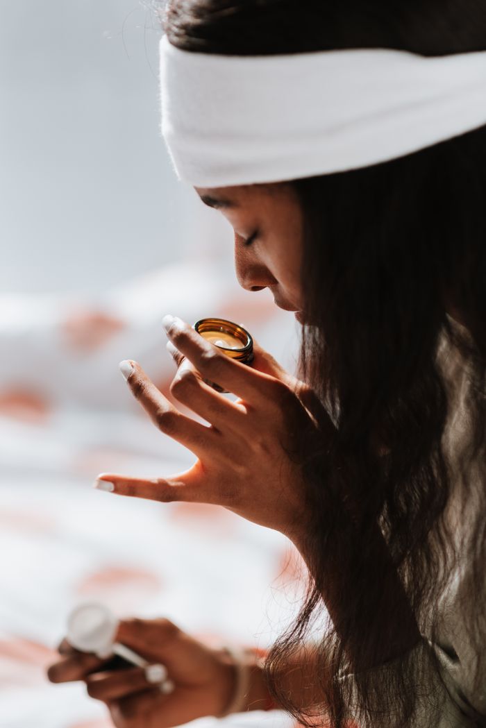 Side view of crop African American female in headband with closed eyes and jar enjoying candle aroma in house